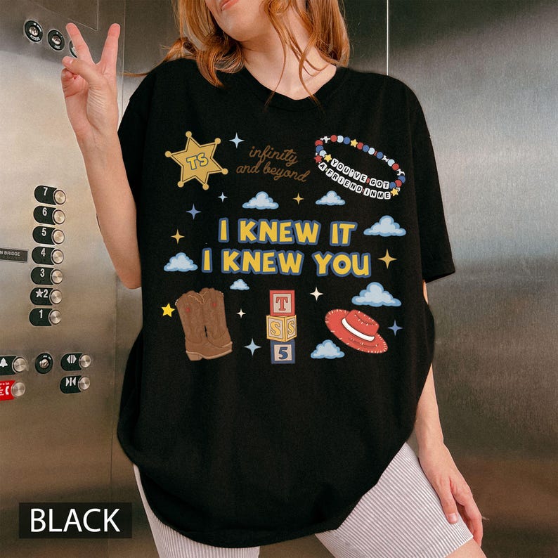 May include: A black t-shirt featuring a graphic print with the text I KNEW IT I KNEW YOU, infinity and beyond, and YOU'VE GOT A FRIEND IN ME. The design includes a gold star, a beaded bracelet, cowboy boots, alphabet blocks, and a red hat.