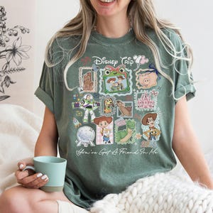 May include: A sage green short-sleeve t-shirt featuring a graphic print with the text "Disney Trip" and "You've Got A Friend In Me". The design includes various Toy Story characters, a castle, and a pig, arranged in a collage of framed images.