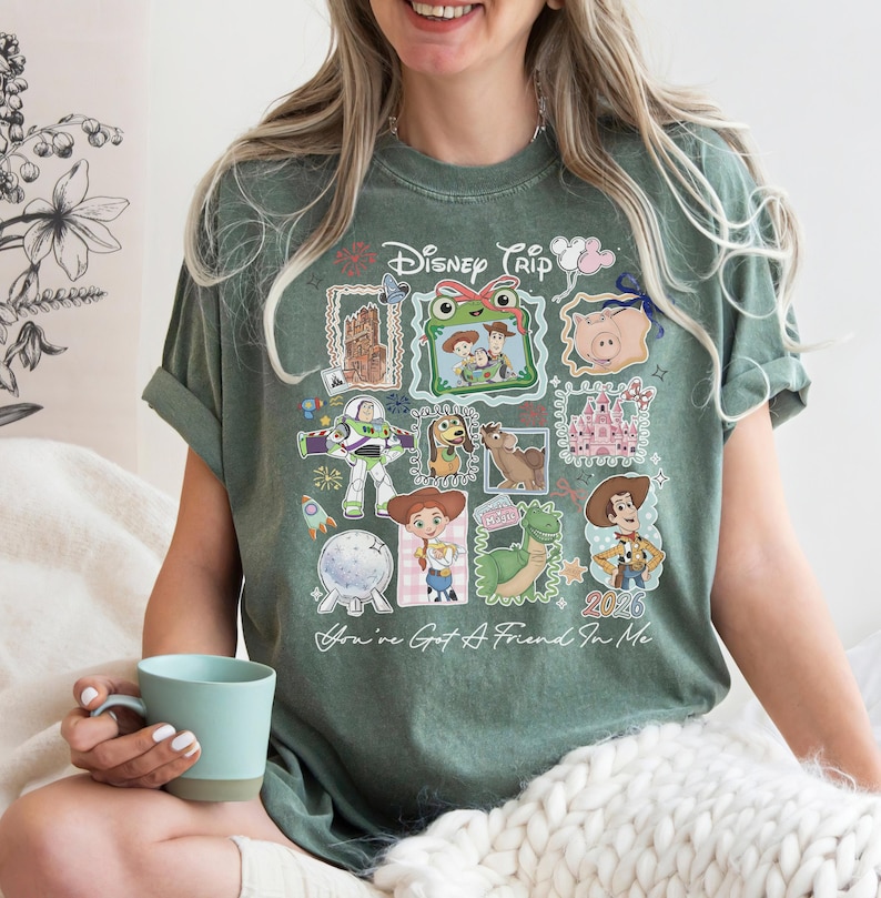 May include: A sage green short-sleeve t-shirt featuring a graphic print with the text "Disney Trip" and "You've Got A Friend In Me". The design includes various Toy Story characters, a castle, and a pig, arranged in a collage of framed images.