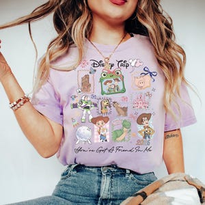 May include: A light purple t-shirt featuring a graphic print with the text Disney Trip and You've Got A Friend In Me. The design includes illustrations of various animated characters, a castle, a rocket, and the year 2026 in a collage style.
