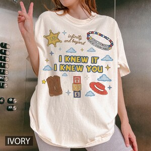 May include: An ivory t-shirt featuring a graphic print with the text I KNEW IT I KNEW YOU, infinity and beyond, and YOU'VE GOT A FRIEND IN ME. The design includes a yellow sheriff star, a beaded bracelet, brown cowboy boots, alphabet blocks, and a red hat.