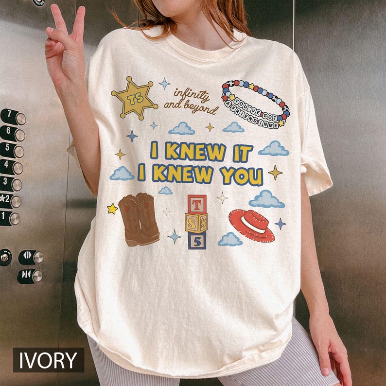 May include: An ivory t-shirt featuring a graphic print with the text I KNEW IT I KNEW YOU, infinity and beyond, and YOU'VE GOT A FRIEND IN ME. The design includes a yellow sheriff star, a beaded bracelet, brown cowboy boots, alphabet blocks, and a red hat.