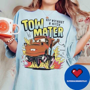 May include: Pale blue t-shirt with a graphic of Tow Mater from the Pixar Cars film. The design includes the text "TOW MATER" and "OFF WITHOUT A HITCH" with flames and the Pixar logo. A white mug with a sun design is held in the image.
