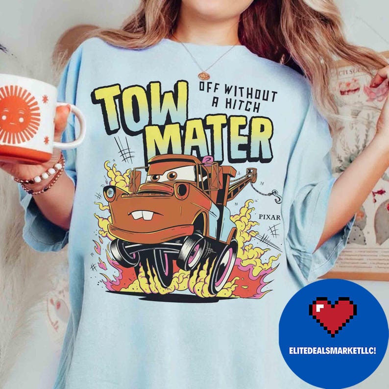 May include: Pale blue t-shirt with a graphic of Tow Mater from the Pixar Cars film. The design includes the text "TOW MATER" and "OFF WITHOUT A HITCH" with flames and the Pixar logo. A white mug with a sun design is held in the image.