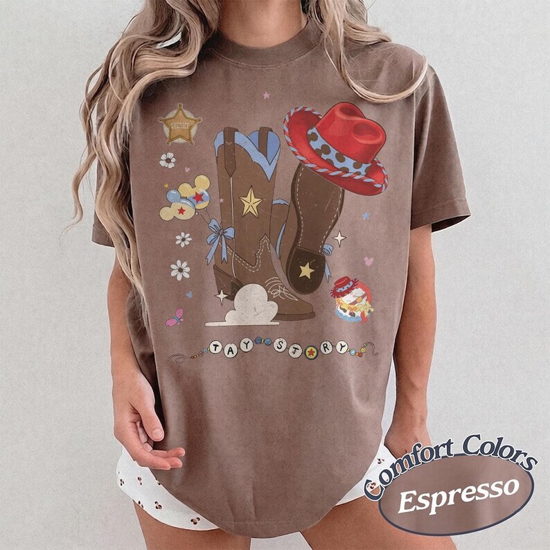 May include: A brown short-sleeve t-shirt featuring a graphic print of brown cowboy boots, a red cowboy hat, a sheriff star, balloons, and small flowers. The text TAY STORY is printed in circular beads at the bottom of the graphic design.