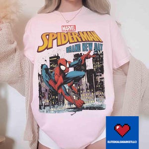 May include: Pale pink t-shirt with the Marvel Spider-Man logo and the text "Brand New Day." The design depicts Spider-Man in action with a city backdrop. The top has short sleeves and a round neck.