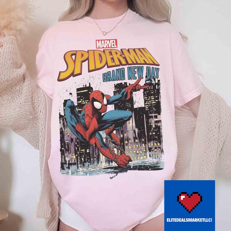 May include: Pale pink t-shirt with the Marvel Spider-Man logo and the text "Brand New Day." The design depicts Spider-Man in action with a city backdrop. The top has short sleeves and a round neck.