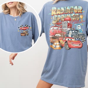 May include: A blue t-shirt featuring a small graphic on the front chest of a brown tow truck with checkered flags and the text Name. The back displays a large graphic of Radiator Springs with various cartoon cars including a red race car.