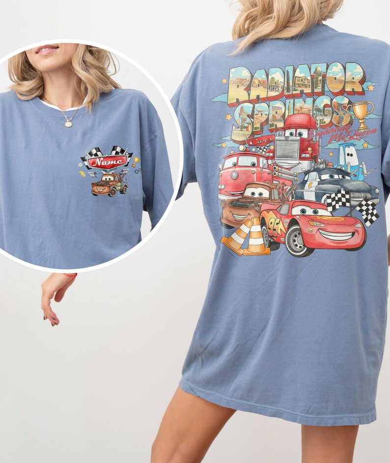May include: A blue t-shirt featuring a small graphic on the front chest of a brown tow truck with checkered flags and the text Name. The back displays a large graphic of Radiator Springs with various cartoon cars including a red race car.