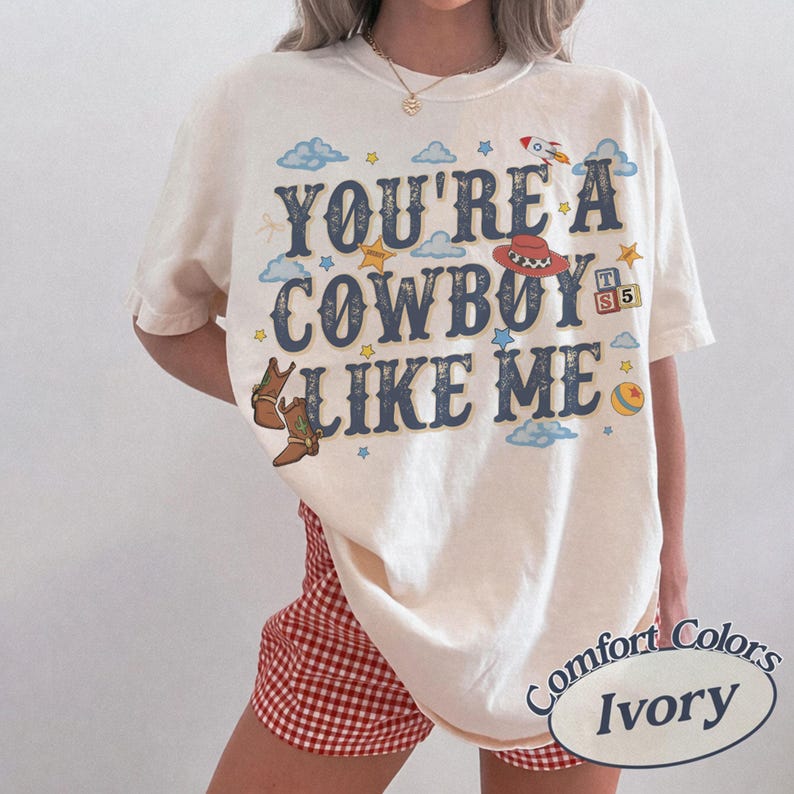 May include: An ivory t-shirt featuring the dark blue text YOU'RE A COWBOY LIKE ME. The graphic includes illustrations of brown cowboy boots, a red cowboy hat, a rocket, a yellow ball, and alphabet blocks with the letters T, S, and 5 among blue clouds.