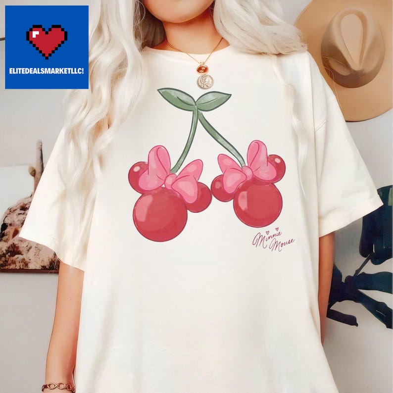 Op de afbeelding: Cr&egrave;mekleurig T-shirt met een grillig ontwerp van twee kersen, elk met een roze strik en groene stelen. De tekst "Minnie Mouse" is geschreven in cursief schrift. Het shirt heeft een relaxte pasvorm.