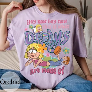 Op de afbeelding: Een lichtpaars T-shirt met een stripfiguur met blond haar en een oranje bloem. De tekst "Hey now hey now, this is what dreams are made of" staat in een retro lettertype. Het woord "Orchid" staat linksonder.