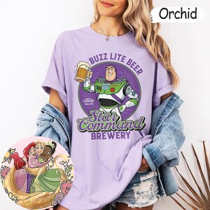 Może przedstawiać: Koszulka w kolorze orchidei z grafiką przedstawiającą Buzza Astrala trzymającego kufel piwa z napisem BUZZ LITE BEER Star Command BREWERY. Okrągła wstawka przedstawia ilustrację Ariel, Roszpunki i Tiany przytulających się do siebie.