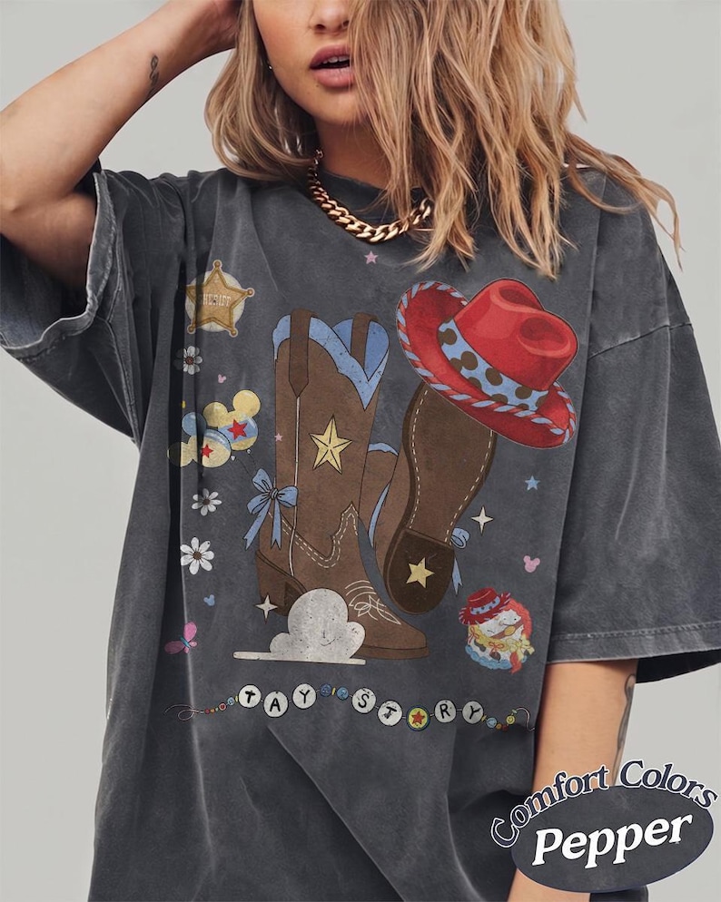 May include: A charcoal gray t-shirt featuring a graphic print of brown cowboy boots, a red cowboy hat, a sheriff badge, flowers, and clouds. The text "TAY STORY" is printed in circular beads at the bottom, with "Comfort Colors Pepper" text.