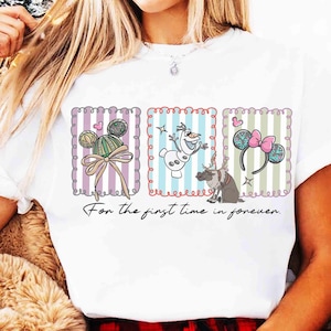 May include: White t-shirt featuring three framed Disney-themed illustrations. The designs include Mickey Mouse ears, Olaf the snowman, and Minnie Mouse ears with a pink bow. The text "For the first time in forever" is written below the illustrations.