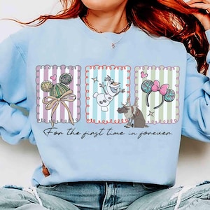 May include: Light blue sweatshirt featuring three framed illustrations. The illustrations include Mickey Mouse ears, Olaf the snowman, and Minnie Mouse ears. The text "For the first time in forever" is printed below the illustrations.