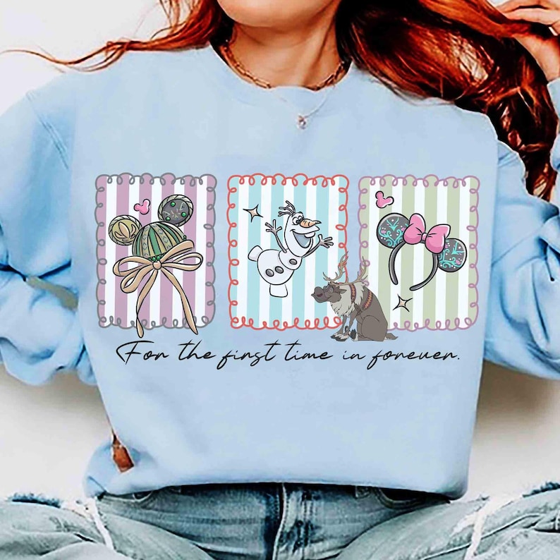 May include: Light blue sweatshirt featuring three framed illustrations. The illustrations include Mickey Mouse ears, Olaf the snowman, and Minnie Mouse ears. The text "For the first time in forever" is printed below the illustrations.