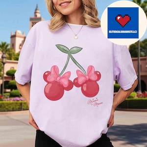 Op de afbeelding: Een lichtpaars T-shirt met een cartoon kers ontwerp met Minnie Mouse strikken en de woorden "Minnie Mouse" in cursief. Het shirt heeft korte mouwen en een relaxte pasvorm. Een blauwe cirkel met een pixelhart op de achtergrond.