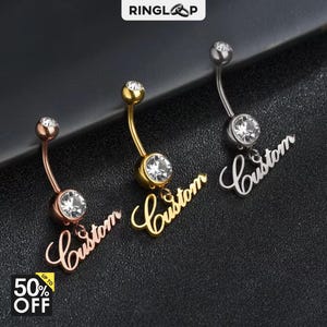 May include: Three belly button rings in rose gold, gold, and silver. Each ring features a curved barbell with a clear gem and a dangling "Custom" script. The image includes the Ringlap logo and a "50% OFF" promotion.