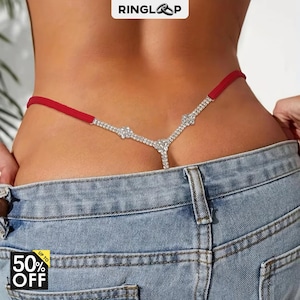 Könnte beinhalten: Ein roter Tanga mit einem Y-förmigen Riemen, der mit klaren Strasssteinen besetzt ist und über einer blauen Jeans getragen wird. Das Design umfasst eine zentrale Strassverzierung und ein schmales rotes Stoffband an Hüfte und Rücken.