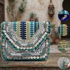 Può includere: Pochette decorata in turchese e argento con chiusura a patta. La borsa presenta un motivo a righe verticali in turchese, blu e bianco. Monete d'argento e pompon turchesi adornano i bordi. Con tracolla a catena argentata. Circa 25 cm di larghezza.