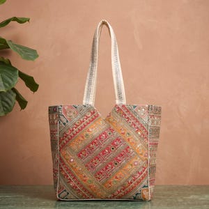 Könnte beinhalten: Eine farbenfrohe Tragetasche mit einem geometrischen Muster in Rot, Orange und Gold. Die Tasche hat dekorative Nähte und Spiegelakzente. Die Griffe sind cremefarben mit einem dezenten Muster.
