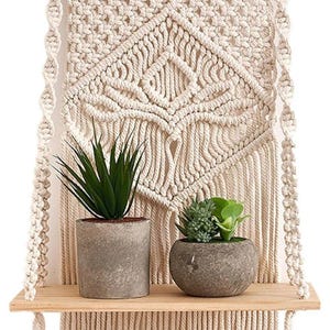Könnte beinhalten: Cremefarbener Makramee-Wandbehang mit einem Holzregal, auf dem zwei Topfpflanzen stehen. Das Makramee hat ein geometrisches Design. Das Regal ist aus hellem Holz und die Töpfe sind grau.