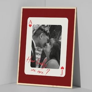Peut inclure: Une photographie encadrée avec une image en noir et blanc d'un couple s'embrassant. La photo est placée sur un fond rouge et encadrée d'une bordure en bois. L'image est superposée d'un motif de carte à jouer, avec l'As de Cœur et le texte "How lucky we are ?".