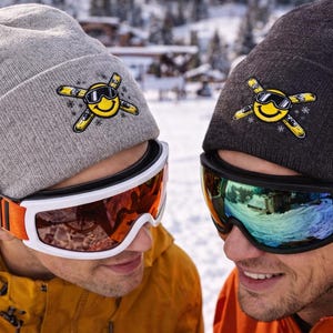 Könnte beinhalten: Zwei Personen tragen Skibrillen und Mützen mit einem gelben Smiley-Logo. Eine Mütze ist grau, die andere dunkelgrau. Die Brillen haben weiße Rahmen und orangefarbene Riemen. Der Hintergrund zeigt eine verschneite Berglandschaft.