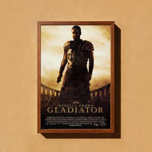 May include: Framed movie poster for the film "Gladiator" starring Russell Crowe. The poster is in a brown wooden frame and shows Crowe in gladiator armour, holding a sword. The film's title, "Gladiator", is clearly visible.