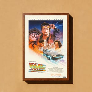 Può includere: Un poster cinematografico incorniciato di "Ritorno al futuro" con una cornice in legno marrone. Il poster presenta un'immagine centrale di un uomo con occhiali da sole, una DeLorean e il testo "Ritorno al futuro" e "40° anniversario".