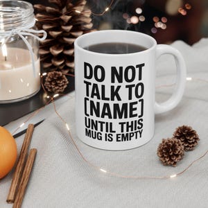 May include: White ceramic mug with black text that reads "DO NOT TALK TO [NAME] UNTIL THIS MUG IS EMPTY." The mug is filled with a dark liquid. The mug is on a light gray surface with decorative items.