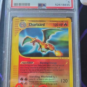Puede incluir: Una carta Charizard Rev. Foil de la edición Pokémon Expedition 2002, calificada. La carta presenta una ilustración de Charizard con una combinación de colores amarillo y naranja. La carta está dentro de un soporte protector con el texto "2002 POKEMON EXPEDITION CHARIZARD - REV.FOIL".