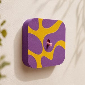 Op de afbeelding: Een vierkante lichtschakelaarafdekking met afgeronde hoeken en een paars en geel golvend abstract patroon. Een kleine, cilindrische lichtroze tuimelschakelaar steekt uit het midden van de plaat, die op een vlak wandoppervlak is gemonteerd.
