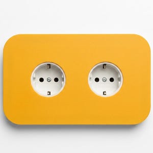 Op de afbeelding: Een rechthoekig dubbel stopcontact met afgeronde hoeken, voorzien van een felgele afdekplaat en twee witte ronde contactpunten met randaarde, ontworpen voor standaard elektrische aansluitingen in huis of op kantoor.