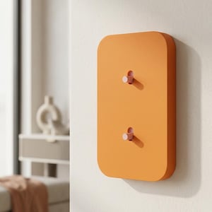 Könnte beinhalten: Eine rechteckige, orangefarbene Wandgarderobe mit abgerundeten Ecken. Das Element verfügt über zwei kleine, hellrosa zylindrische Haken, die aus vertikalen Schlitzen herausragen und eine funktionale Aufbewahrungslösung zum Aufhängen bieten.