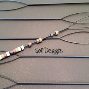 May include: Five black beading tools with a loop at one end and a pointed end. One tool has a string of brown, white, and gold beads threaded through it. The text "Sol Doggie" is visible on the surface.