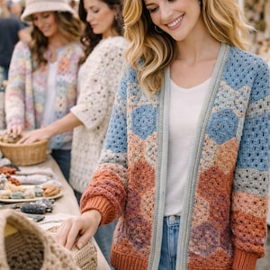 May include: A woman wearing a handmade crochet cardigan in shades of blue, orange, and peach. The open-front sweater features a geometric pattern and is paired with blue jeans. Other women are seen wearing similar crochet garments.