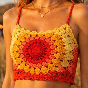 Op de afbeelding: Een kleurrijke gehaakte top met een vierkante halslijn en dunne bandjes. De top heeft een cirkelvormig patroon in rood, oranje en geel, met een geschulpte rand. Een levendig handgemaakt kledingstuk.