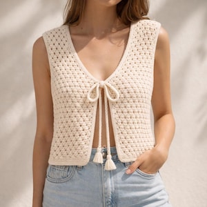 Puede incluir: Chaleco de crochet color crema con cuello en V y cierre de lazo. El chaleco sin mangas presenta un diseño de tejido abierto texturizado y pequeñas borlas en la parte inferior de los lazos. Una prenda elegante hecha a mano.