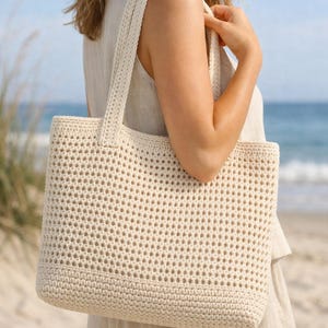 May include: A cream-coloured, open-weave crochet tote bag with strong handles. The bag has a rectangular shape and a textured appearance. The bag is being held by a person on a beach, suggesting a casual, summery style. The bag is a neutral colour.