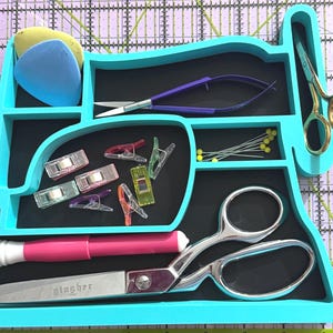 May include: A turquoise sewing machine-shaped organizer filled with sewing tools. Includes scissors, pins, clips, and chalk. The organizer has a black interior and is set on a cutting mat.