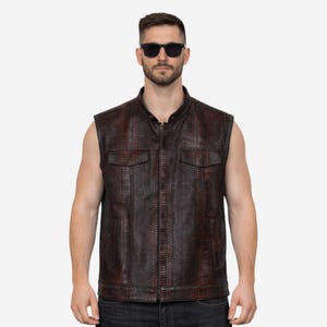 May include: A sleeveless brown leather vest with a textured, snake-skin pattern. The vest has a zip-up front, two chest pockets, and a stand-up collar. The person in the image is wearing black sunglasses and black jeans.