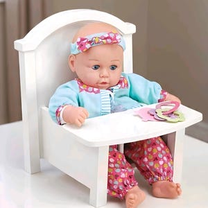 Peut inclure: Une poupée assise dans une chaise haute en bois blanche. Elle porte une tenue bleue avec des détails roses et un pantalon à motifs. La poupée a un bandeau assorti et tient des formes de jouets colorées.