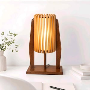 Puede incluir: Una lámpara de mesa con una pantalla cilíndrica hecha de listones de madera clara verticales que rodean una fuente de luz cálida. La lámpara está sostenida por un marco de madera marrón oscuro con dos pilares laterales y una base rectangular.