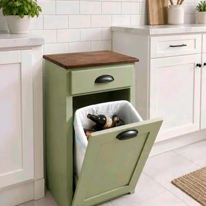 May include: A sage green wooden cabinet with a dark brown top and a drawer. The cabinet door is open, revealing a white bag holding wine bottles. Black handles are on the drawer and door. The cabinet is in a kitchen setting.