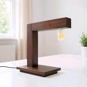 May include: A dark brown wooden desk lamp with a rectangular base and a vertical support. The lamp has a horizontal arm with a lightbulb hanging from it. The lightbulb has a warm, yellow glow. The lamp is on a white surface.