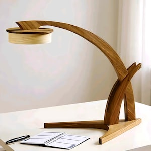 May include: A contemporary wooden desk lamp with a curved arm and a circular light fixture. The lamp is made of light and dark wood tones and sits on a white desk. A notebook and pen are also on the desk.