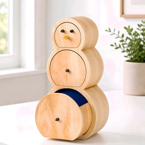 May include: A wooden snowman-shaped cabinet with three drawers. The cabinet is made of light-coloured wood and features black knobs and a small yellow beak. The bottom drawer is open, revealing a blue interior.