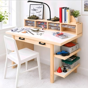 Puede incluir: Un escritorio de manualidades de madera clara con superficie de trabajo blanca, que incluye un estante con cajones transparentes, una lámpara de escritorio negra y dos tiradores negros. Cuenta con estantes laterales y una silla blanca.